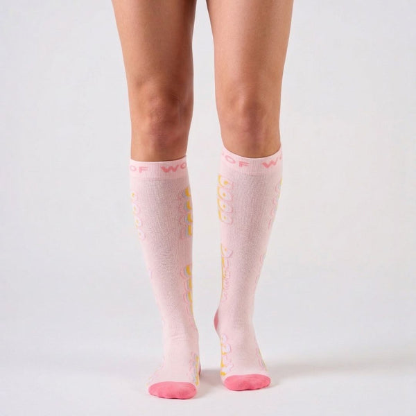 Studio shot of a pair of light pastel pink Good Vibes Bamboo Compression Socks worn on leg models, featuring vertical yellow typography graphics reading "GOOD VIBES", bright pink contrast toe caps and heels, and a matching pink woven top cuff.   