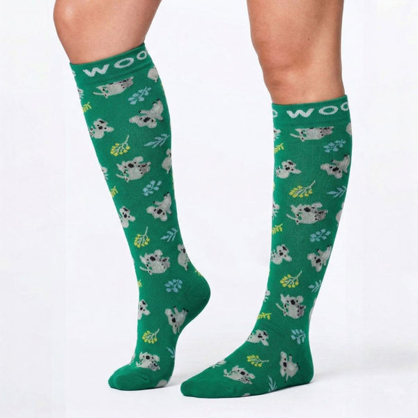 Koalas Bamboo Compression Socks worn on leg models, featuring a repeating pattern of fluffy grey koalas climbing and clinging to eucalyptus branches with yellow blossoms, finished with matching green heels and toes and a minty green "WOOF" woven top cuff.