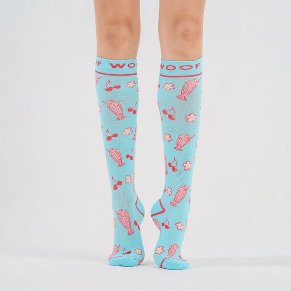 Nurse wearing light blue knee-high compression socks printed with pink manatee illustrations and pink top cuff branding.